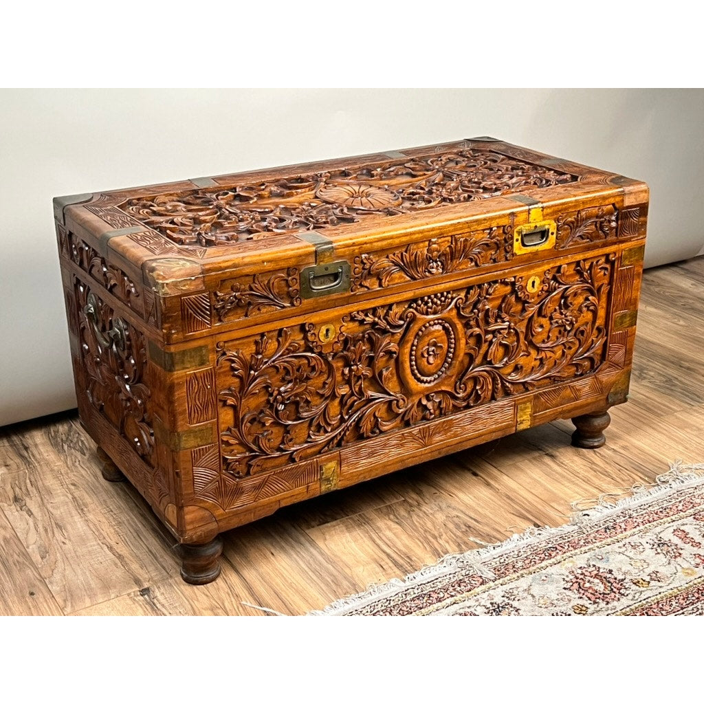 A Chinese Export Carved Camphor Wood Dowry Chest, featuring intricate floral and geometric carvings, metal latches, and short legs, sits on a wooden floor beside a patterned rug.