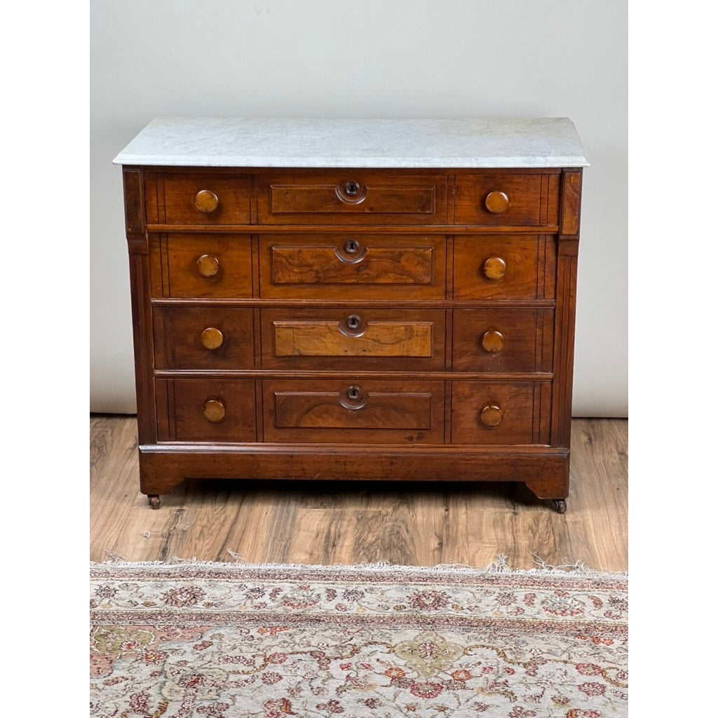 An Antique Victorian Marble Top Four Drawer Chest c. 1910 stands on a wood floor next to a patterned area rug.