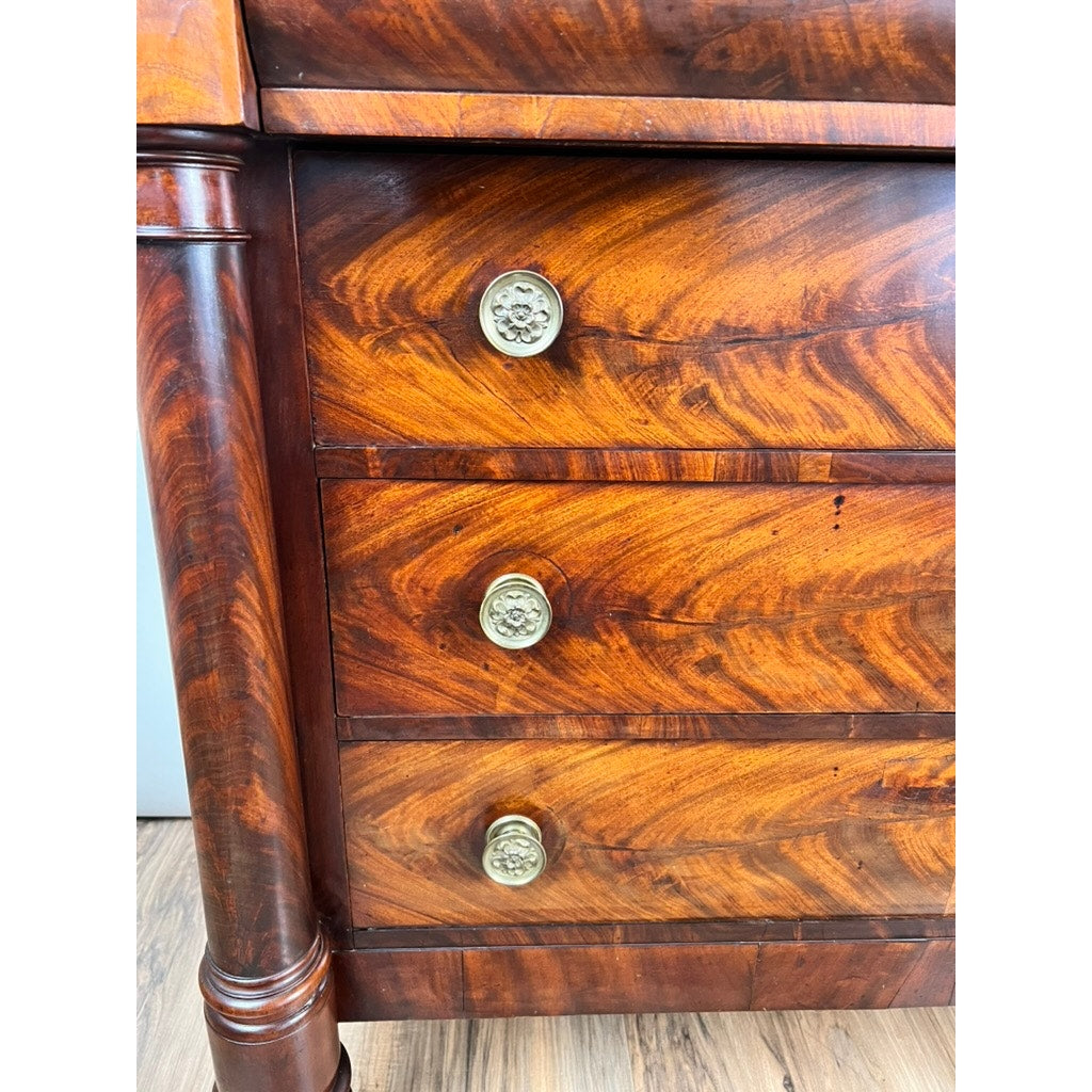 Close-up of an Antique American Empire Albany Four Drawer Chest c. 1840 with ornate brass knobs, decorative carved legs, and rich flame mahogany grain. The chest stands on a light-colored wood floor. Brand: Unknown.