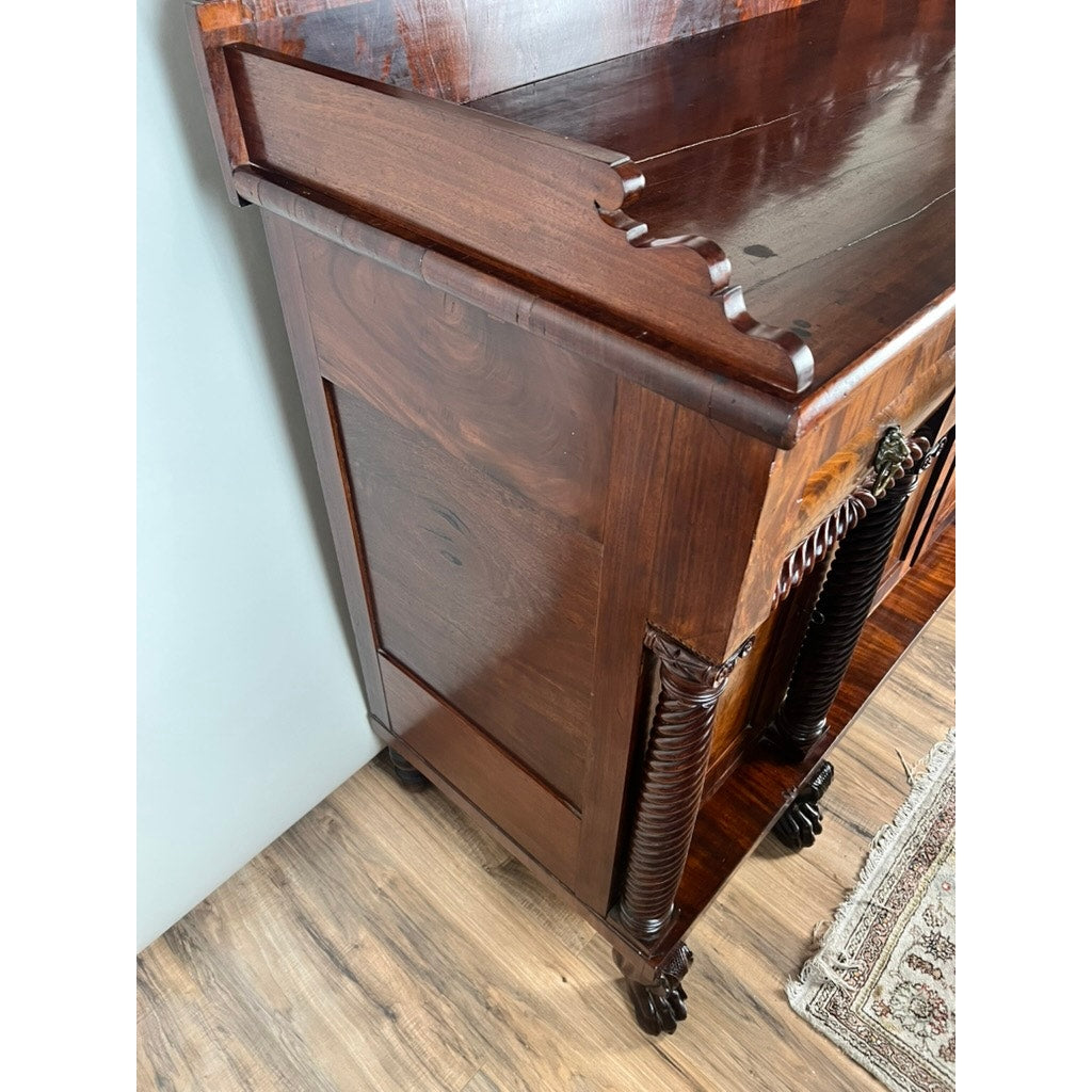 A detailed view of an Antique American Empire Mahogany with Rosewood Edge Banding Sideboard, likely from Philadelphia c.1840, featuring carved legs and ornate details, placed on a wooden floor near a light wall and patterned rug.