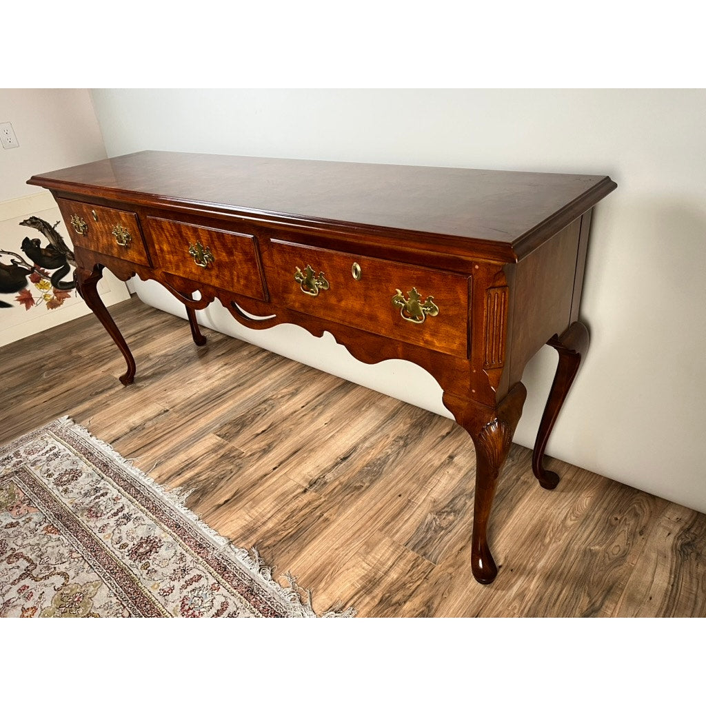 A Vintage Solid Cherry Queen Anne Style Sideboard by Henredon stands polished against a light wall on a wood floor beside a patterned rug, featuring ornate brass handles, cabriole legs, and three drawers.