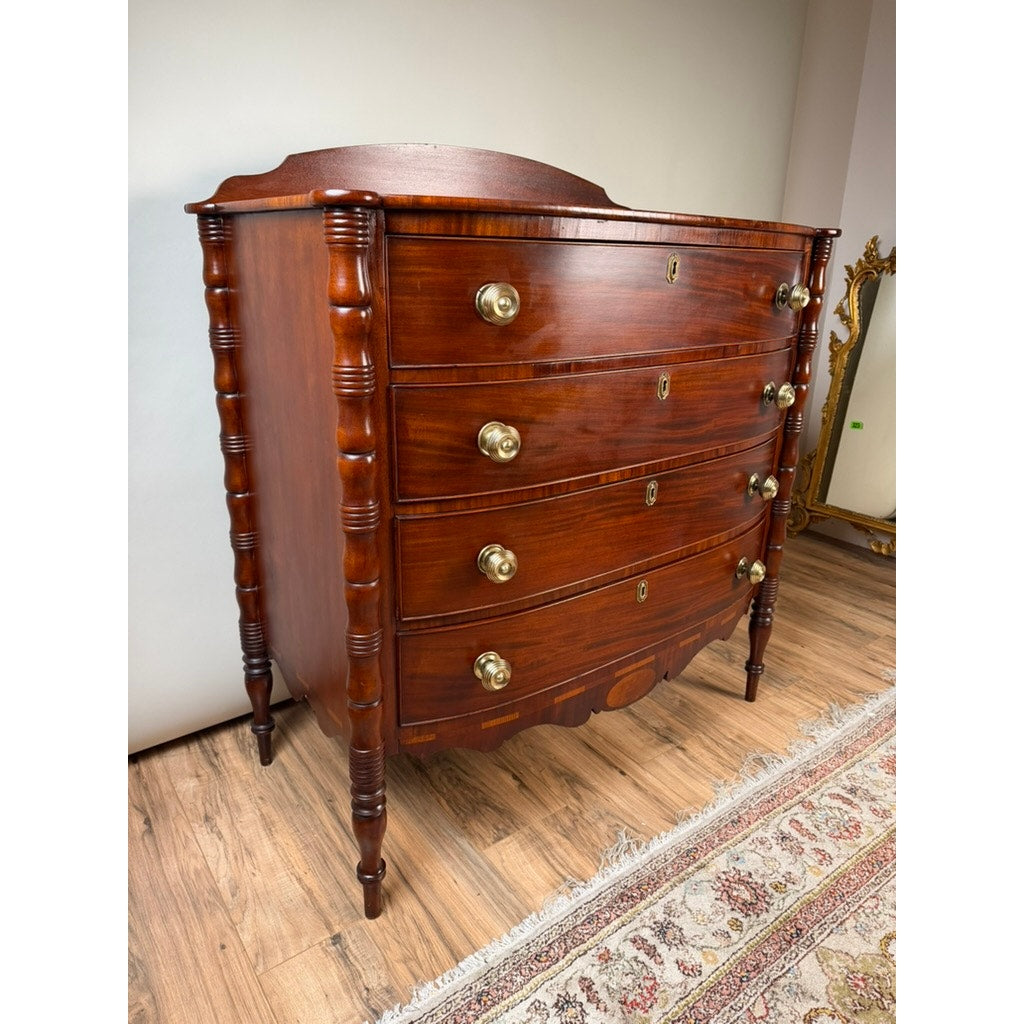 An Early 19th Century New England Federal Mahogany Bow-Front Chest of Drawers with Backsplash, c. 1820–1840, stands on wooden flooring beside a patterned rug and a decorative mirror. This elegant antique piece features four gracefully curved drawers adorned with brass knobs and decorative carved columns on the sides. Brand: None and/or Unknown.