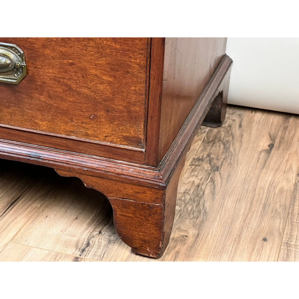 Close-up of the corner of a Late Georgian English Mahogany Chest of Drawers, Circa 1780–1810, featuring brass hardware and standing on a wood-patterned floor. The period chest displays some wear on its legs and edges.