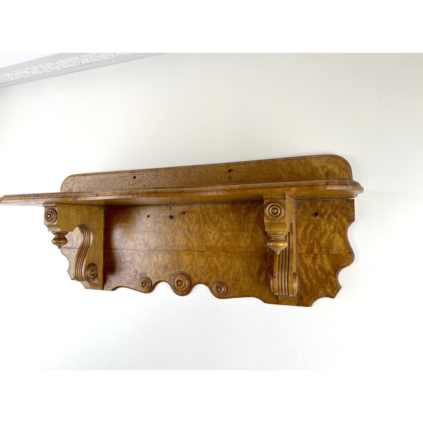 The Antique Victorian Birdseye Maple Wall Shelf c. Late 19th Century features ornate carved details, scrollwork, decorative supports, and a wavy bottom edge, shown mounted on a white wall for timeless elegance. Brand: Unknown.
