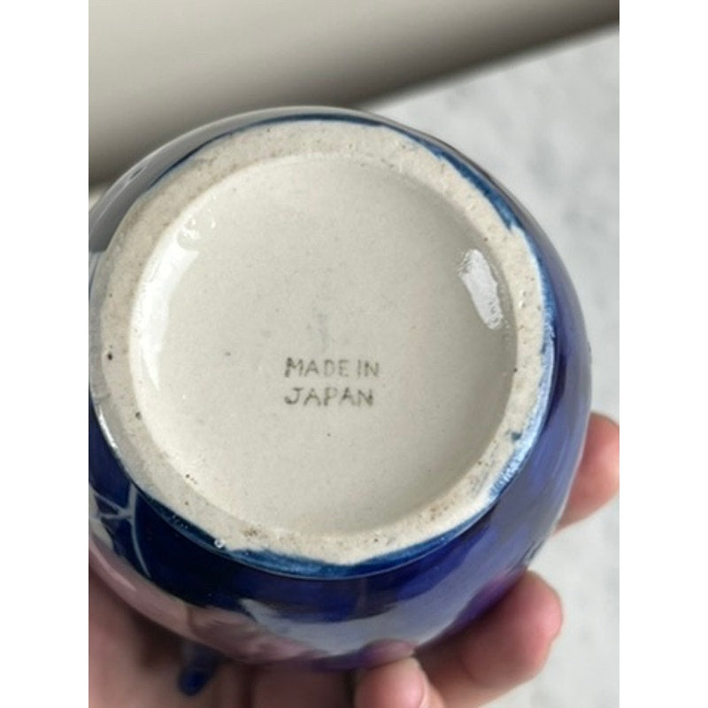 A hand holds a Vintage Japanese Blue and White Floral Bud Vase with a glossy finish and white base marked “MADE IN JAPAN.” Its blue-and-white design reflects light indoors, showcasing classic floral pottery craftsmanship. Brand: Unknown.