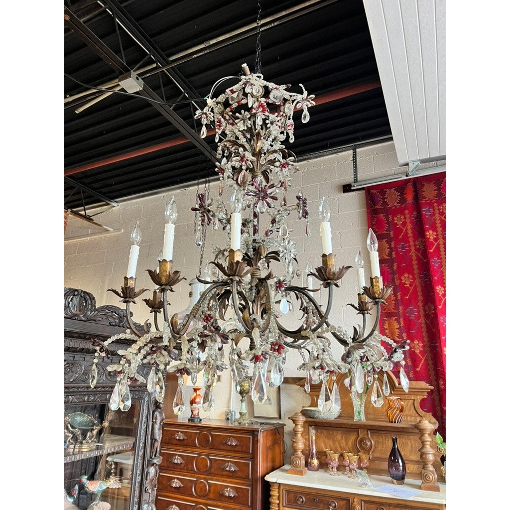 An Antique Late 19th Century French Rococo Tole Amethyst and Crystal Ten Light Chandelier hangs from a black ceiling above antique wooden furniture, illuminating a vintage room with red patterned curtains.