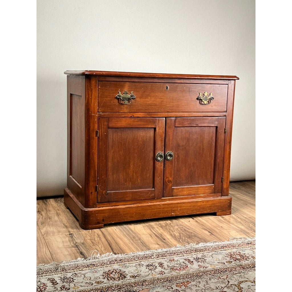 The Antique Victorian Pine Commode c. 1900, with a top drawer, two front doors, round metal handles, and brass hardware, sits on a wooden floor by a rug and light wall—ideal for stylish bedroom storage.