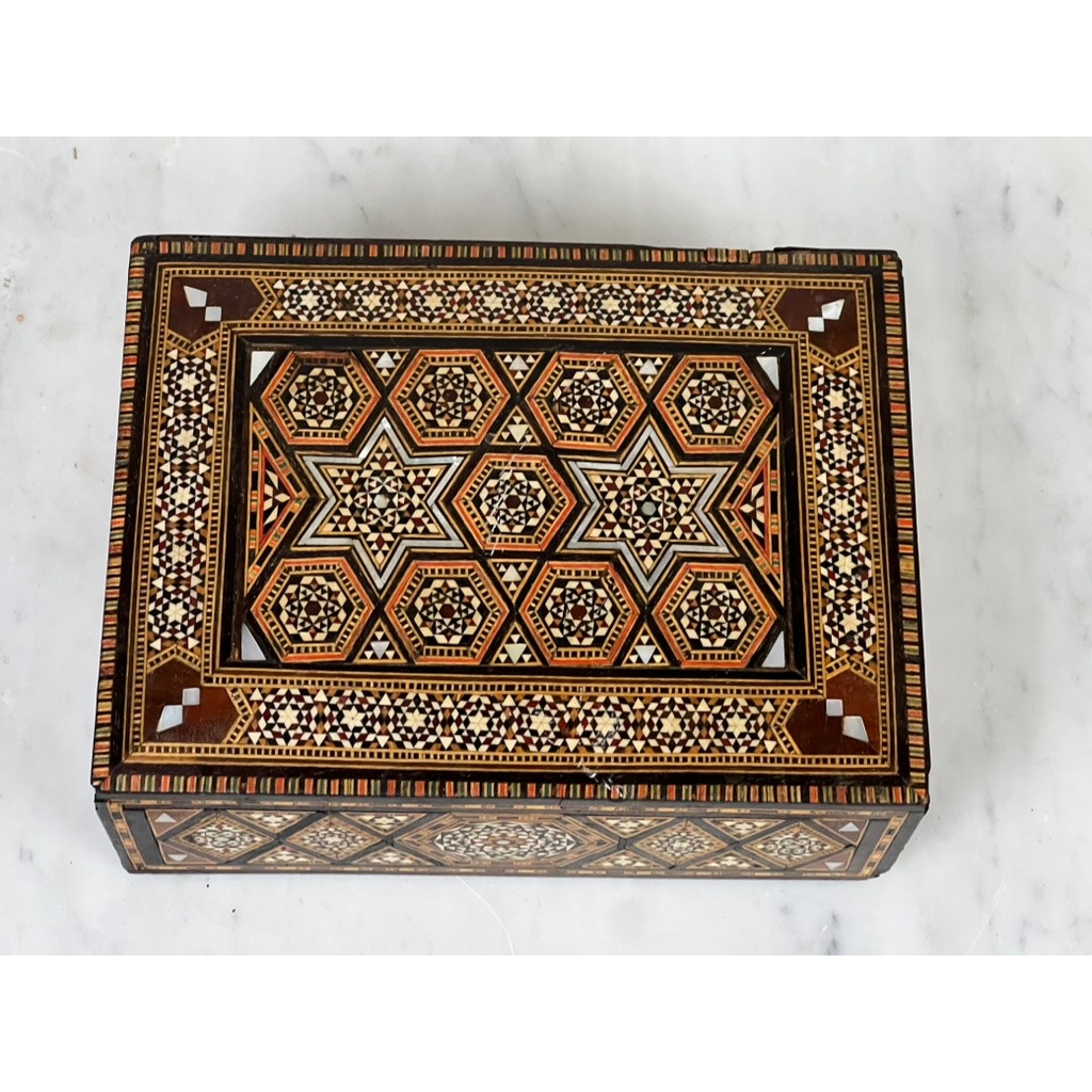 An Antique Persian (probably Syrian) Khatam inlaid wood secret drawer jewelry box from the late 19th century, featuring star-shaped mosaic patterns in brown, gold, black, and white, rests on a white marble surface.