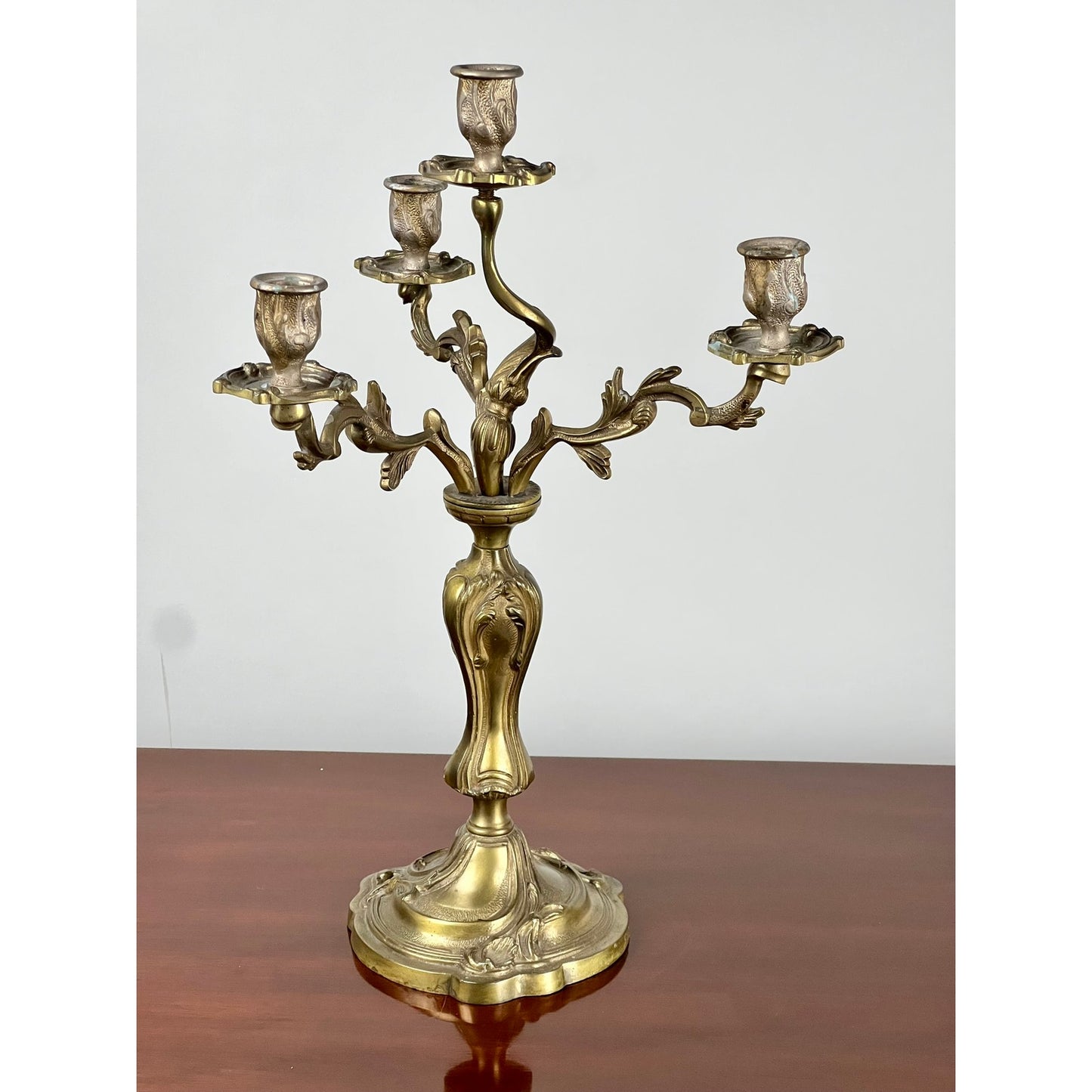 A Vintage Brass Louis XV Style Four Light Candelabra, featuring intricate leaf designs, stands on a polished wooden surface against a plain, light-colored background. Brand: None and/or Unknown.