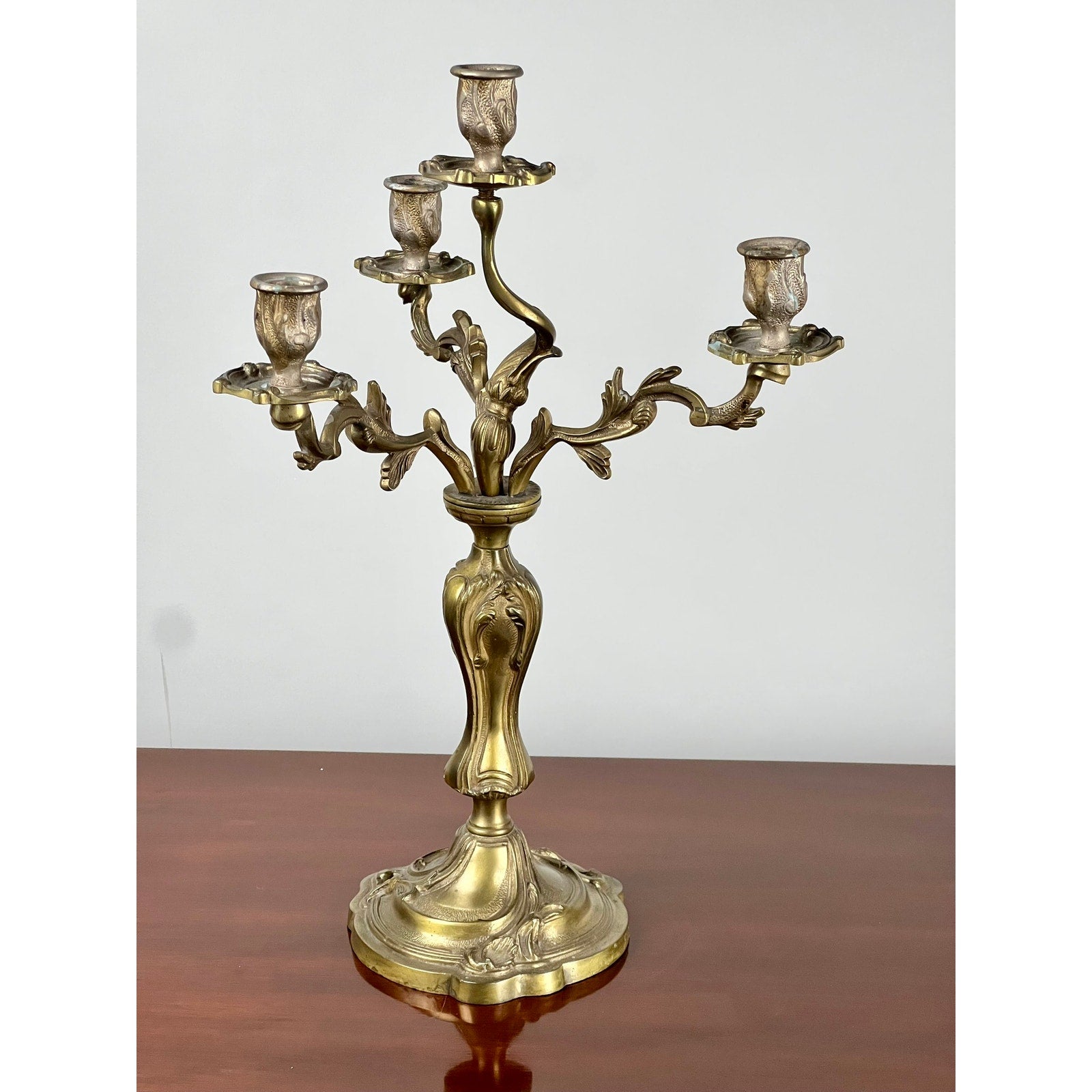 A Vintage Brass Louis XV Style Four Light Candelabra, featuring intricate leaf designs, stands on a polished wooden surface against a plain, light-colored background. Brand: None and/or Unknown.