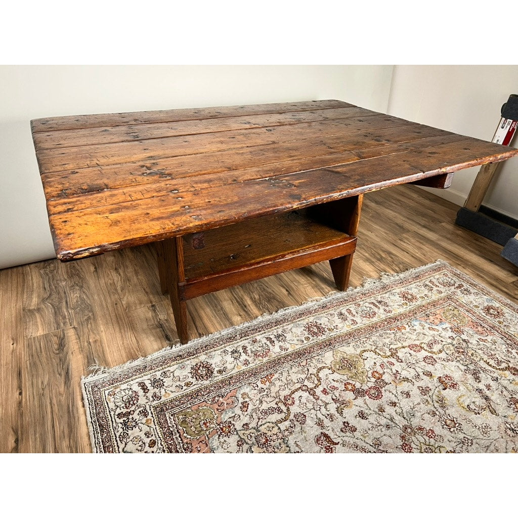 An Antique Country Pine Hutch Table with Six Plank Top c. 18th Century, featuring a weathered finish and lower shelf, stands on a wood floor beside a patterned rug in a simply decorated room with light-colored walls.