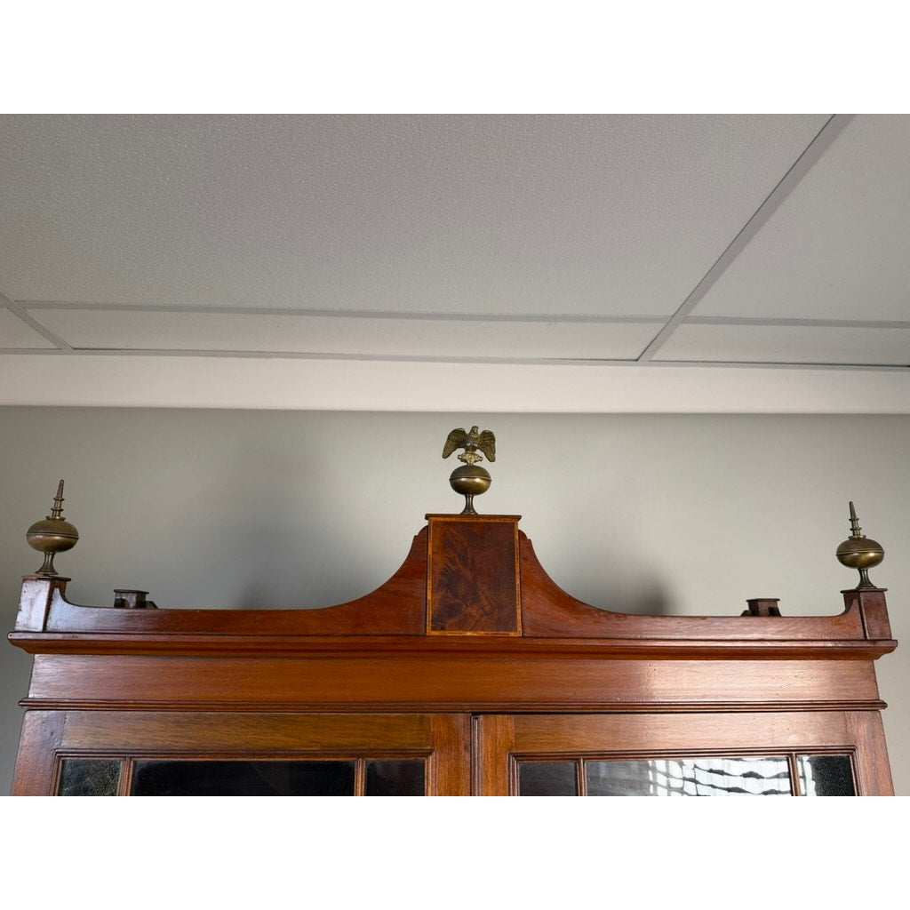 The top section of the Federal Flame Mahogany Secretary Bookcase with Eagle Finial, c. 1810–1825, displays exquisite early 19th-century American cabinetmaking in richly figured flame mahogany, and features a decorative brass eagle finial against a light gray wall and a white tiled ceiling.