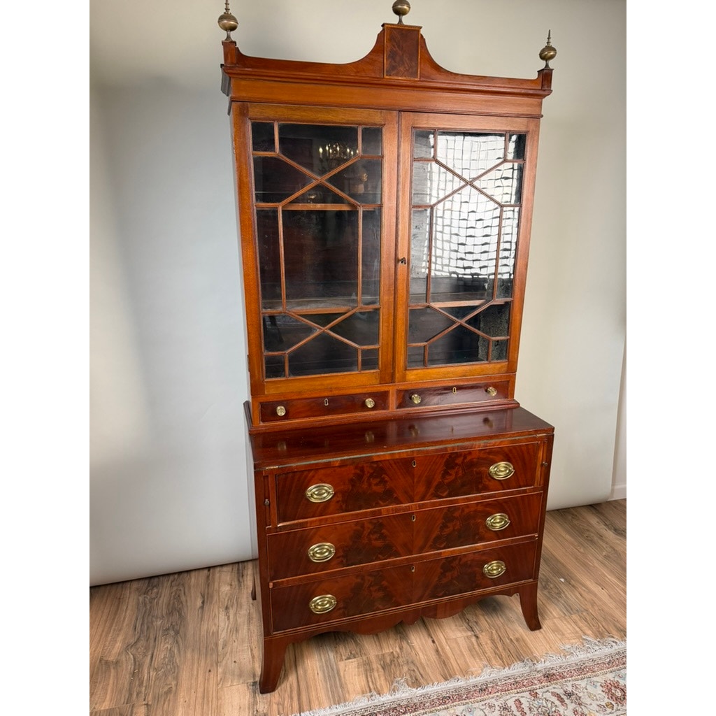 Federal Flame Mahogany Secretary Bookcase with Eagle Finial, c. 1810–1825, featuring tall flame mahogany construction with glass-paneled doors on the upper section, four lower drawers adorned with decorative brass handles, and intricate woodwork characteristic of early 19th-century American Federal style. This period secretary bookcase stands on short legs and is shown placed on a hardwood floor beside a patterned rug.