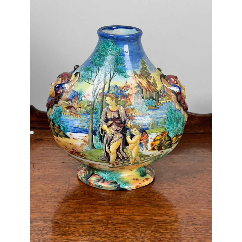 An Antique Italian Renaissance Revival Hand Painted Maiolica Vase (c. 1920-1940) with vibrant painted scenes and winged putti handles is displayed on a wooden surface. Brand: Unknown.