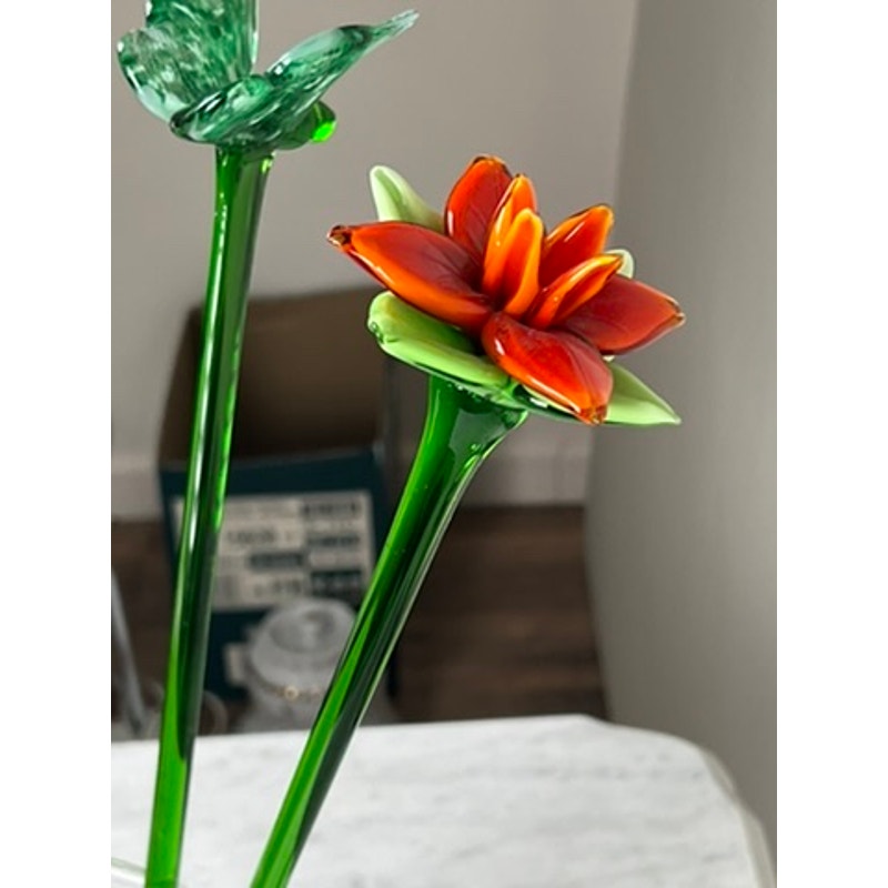 The Vintage Bouquet Murano Art Glass Long Stem Flowers and Butterflies features two hand-blown glass flowers—one with red-orange petals, the other green—each with green stems, displayed on a white surface with a softly blurred background.