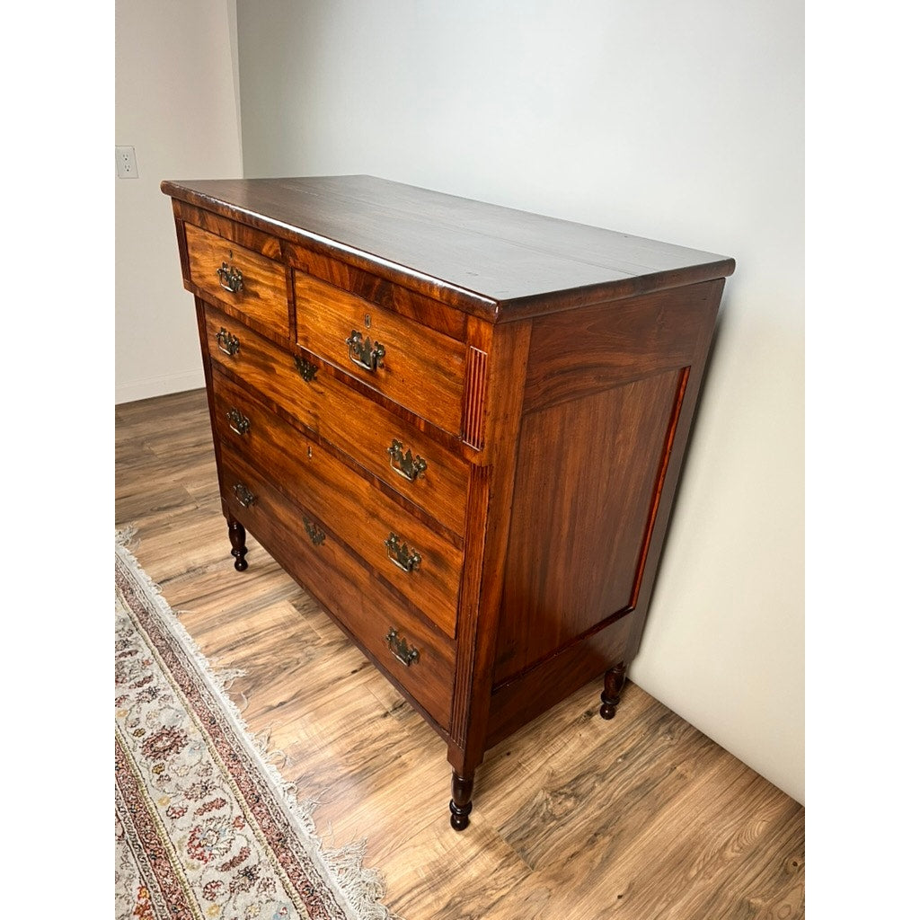 An Antique George III Sheraton Flame Mahogany Chest of Drawers c. Early 19th Century, featuring six drawers and brass handles, stands on a wood floor beside a patterned rug and a plain light-colored wall.