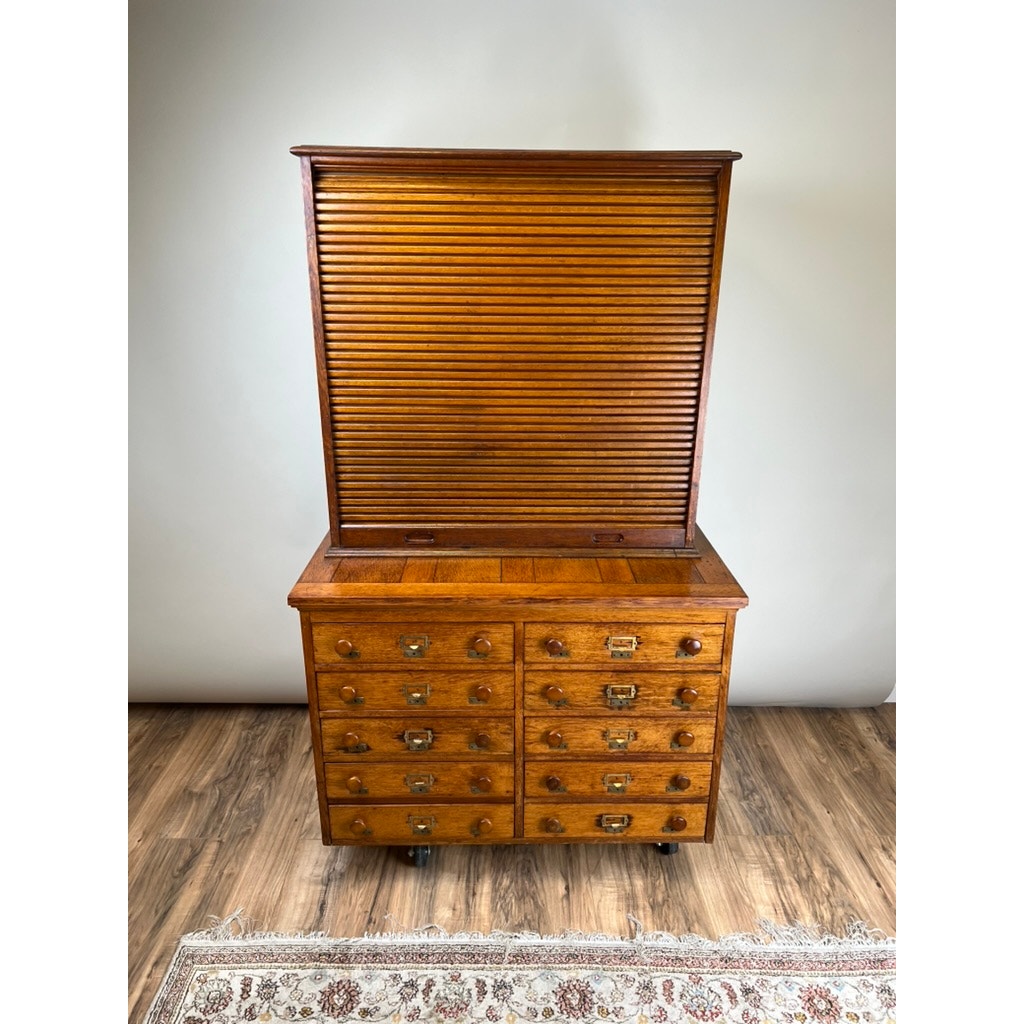 An Antique Early 20th Century Oak Multidrawer File with Roll Top Pigeon Hole Top sits on a hardwood floor, its roll-top cover pulled down and a decorative rug partially visible in front; brand is unknown.