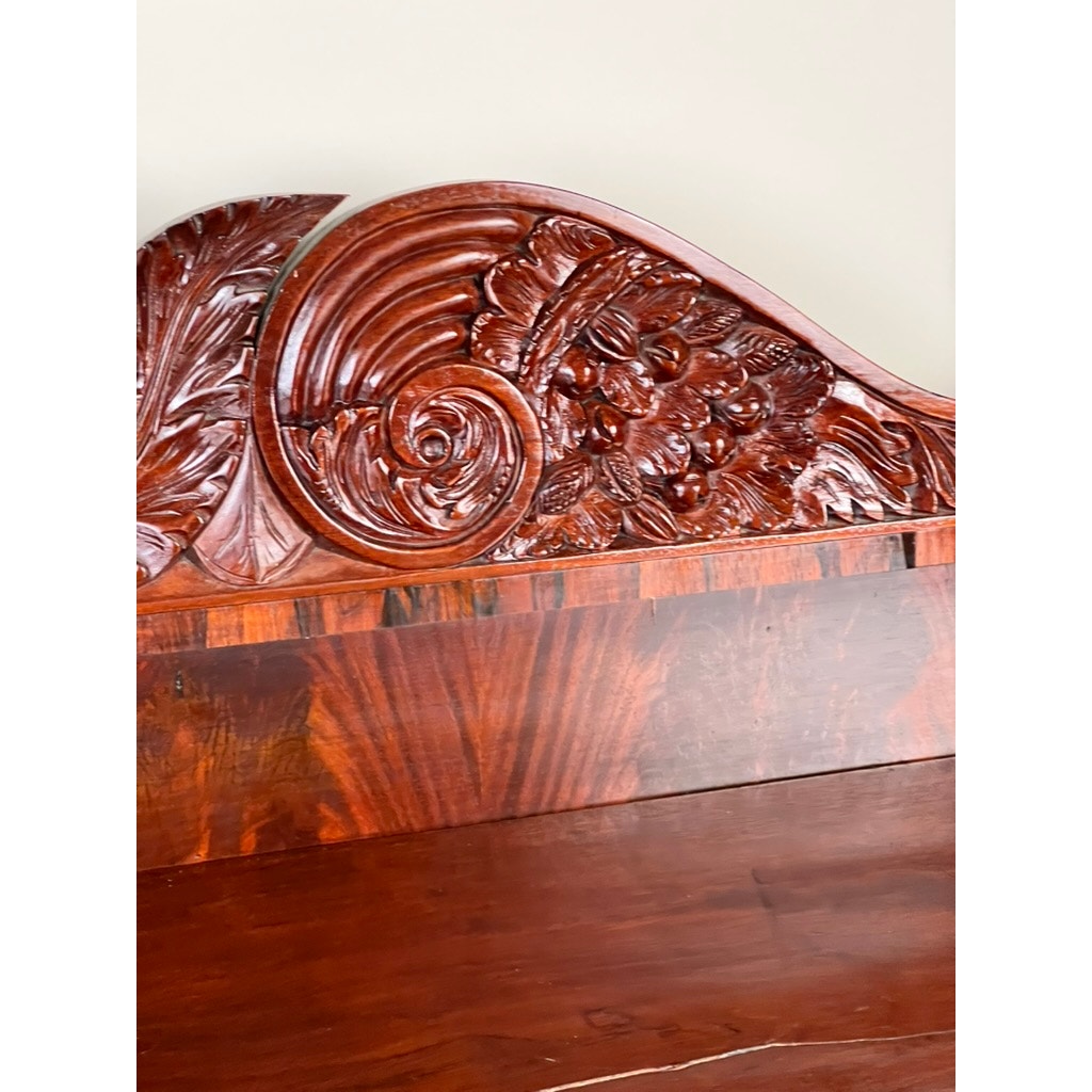 Close-up of an Antique American Empire Mahogany Sideboard, probably Philadelphia c. 1840, with rosewood edge banding—featuring ornate carved designs, swirling patterns, and floral and leaf motifs set against a plain light background.