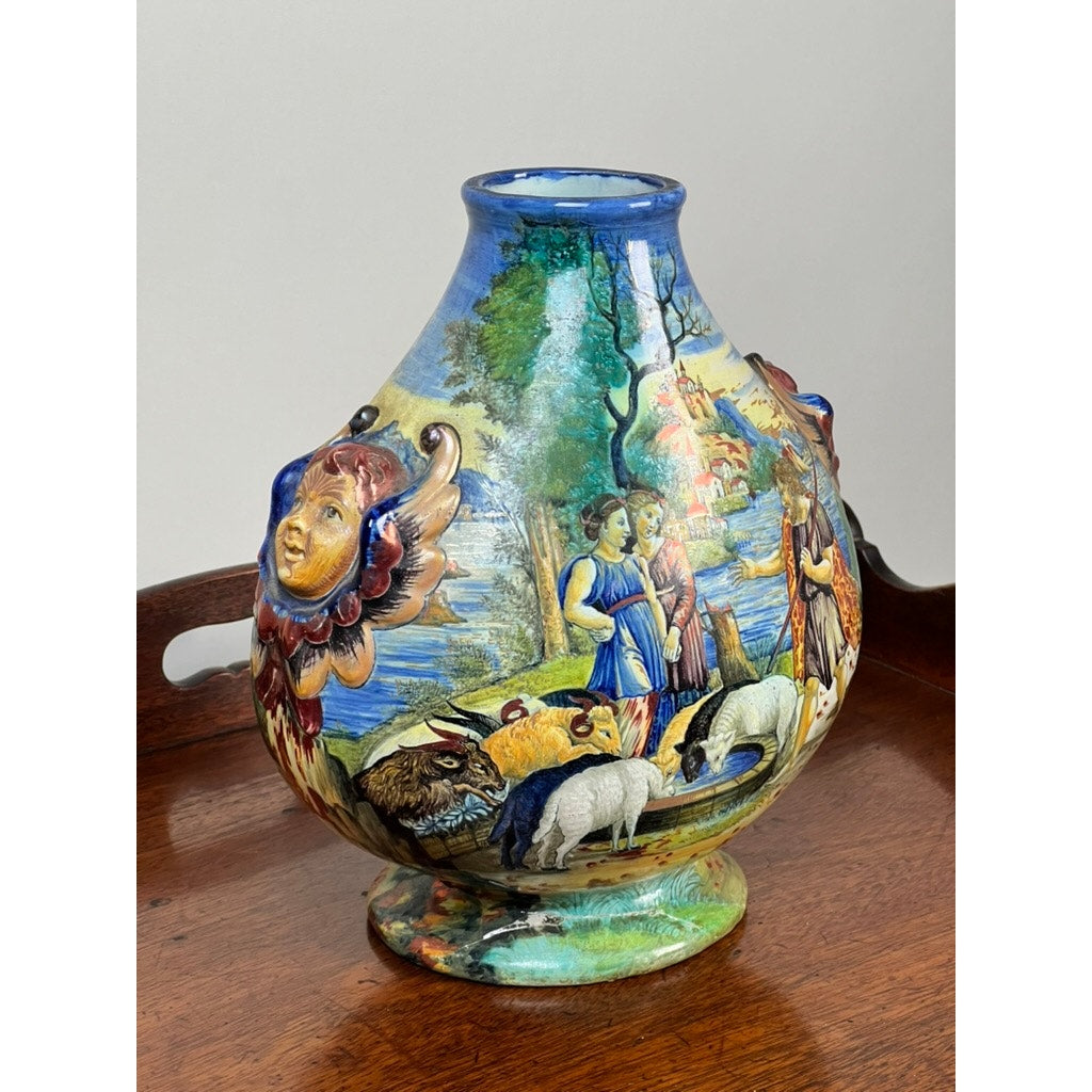 Antique Italian Renaissance Revival maiolica vase, c. 1920-1940, hand painted with pastoral scenes and sculpted faces; features winged putti handles and sits on a wooden surface. Brand: unknown.