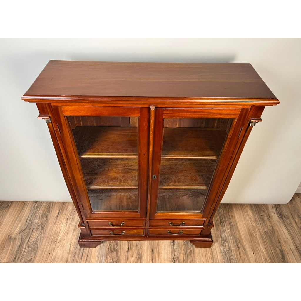 The Vintage Victorian Style Glass Door Mahogany Bookcase stands on a wood floor, featuring elegant glass doors, two interior shelves for display, and four bottom drawers for storage. Brand: Unknown.