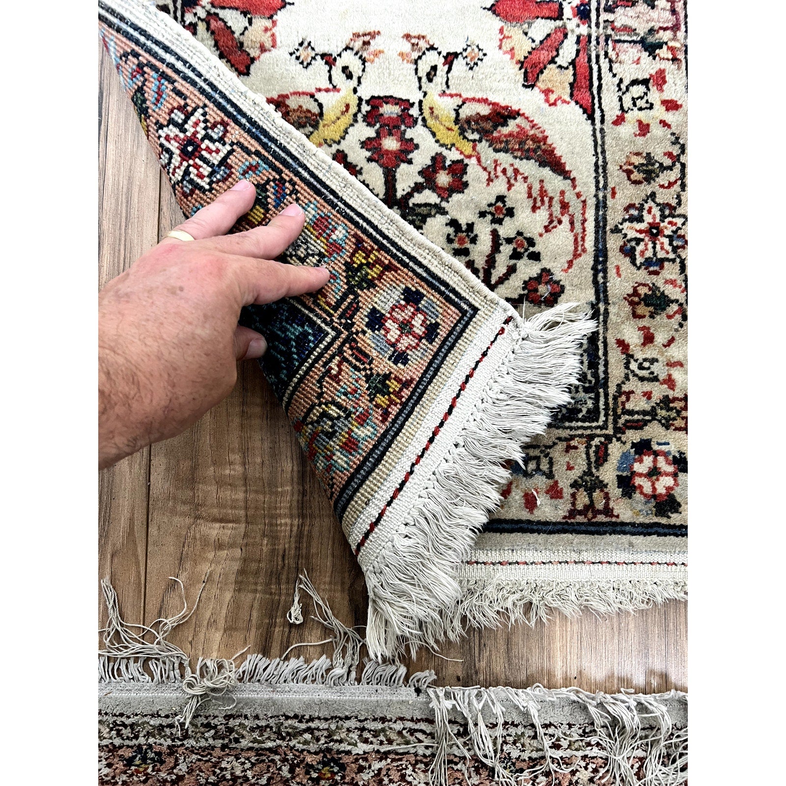 A hand lifts the corner of a Vintage Pair of Hand-Knotted Oriental Tree of Life Scatter Rug (1.5 x 2.5), showing detailed weaving and a bird/floral design. The rug rests on wood flooring with another vintage rug partly visible below.