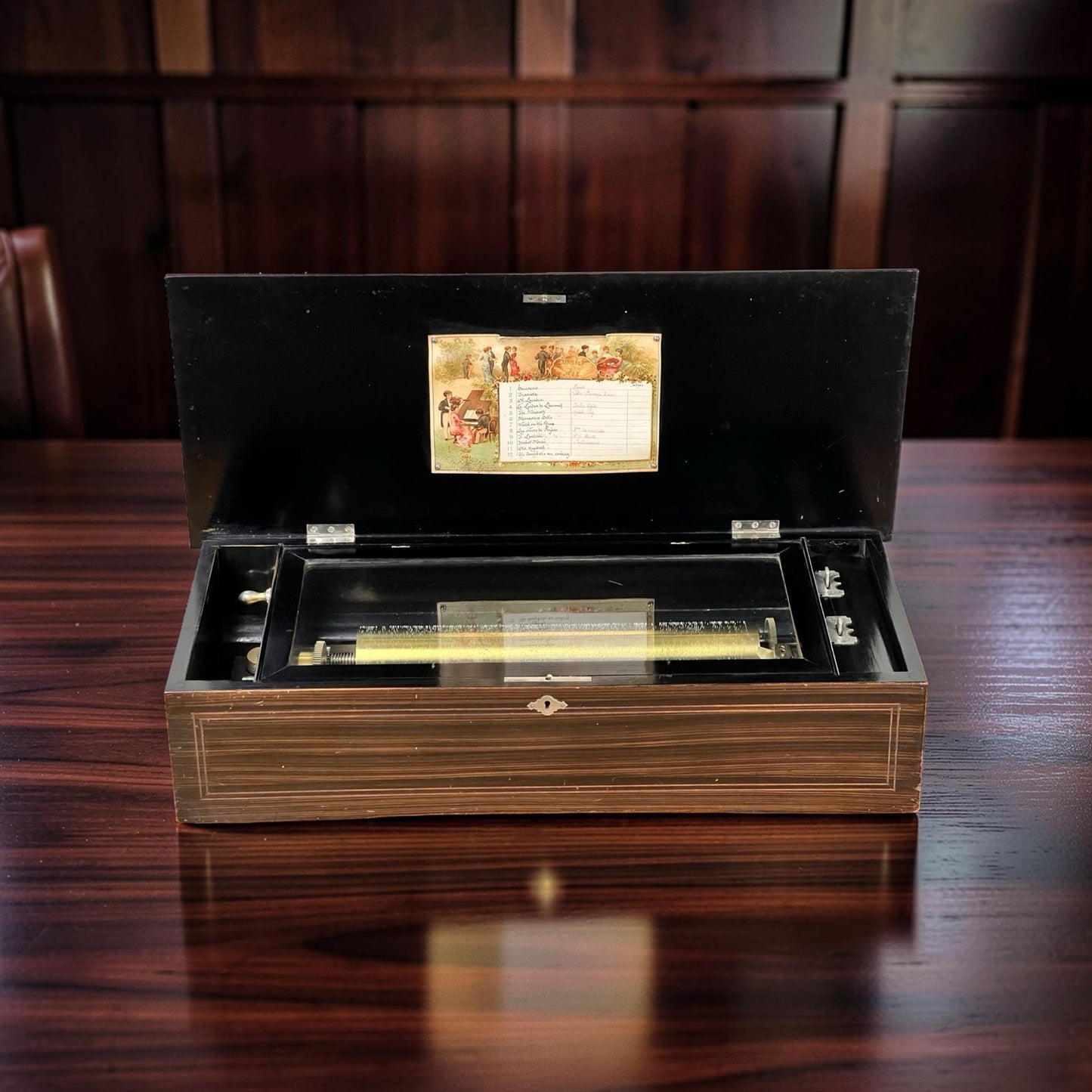 An Antique Swiss 12 Tune Music Box c. Late 19th Century, with an open wooden case revealing its inner mechanism and a decorative label in the lid, sits on a polished wood table before wood-paneled walls. Brand: None/Unknown.