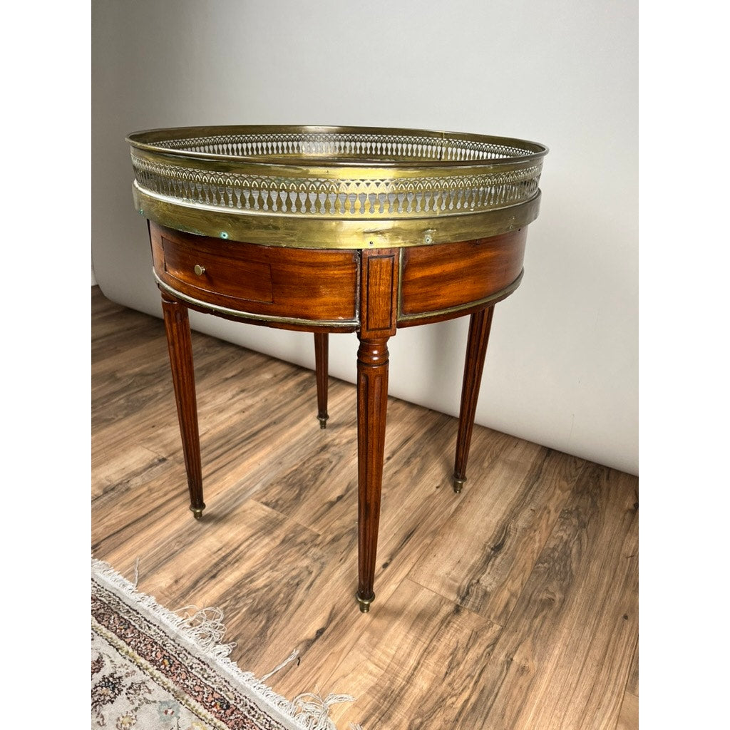 An Antique 19th Century French Louis XVI-style mahogany bouillotte table with marble top and pierced brass gallery stands on a wood floor next to a fringed rug. Brand: Unknown.
