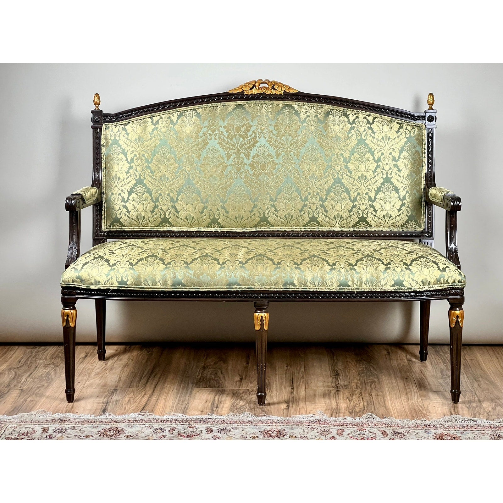 A Vintage Louis XVI Style Settee in Green Damask Upholstery with ornate carvings, decorative armrests, and gold accents is placed on a wooden floor with part of a patterned rug visible. Brand: None/Unknown.