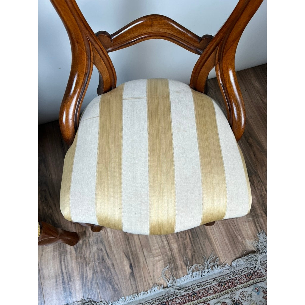 A pair of antique Victorian balloon back chairs from the late 19th century, featuring curved backrests and cream-and-gold striped padded seats, stand on a wooden floor next to a patterned rug.