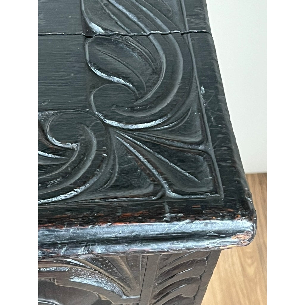 Close-up of the corner of an Antique 18th Century Charles II Style Mule Chest, possibly Irish or Scottish, showing intricate carved leaf patterns, aged dark wood with some wear and a crack, against a light wooden floor. Brand unknown.