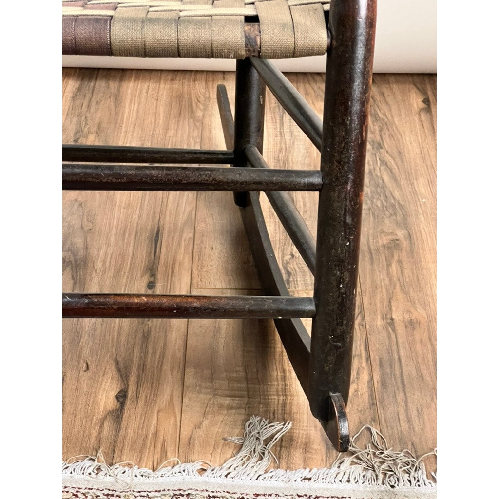 Close-up of the Antique Shaker #6 Tape Seat Rocker by Mt. Lebanon Shakers, late 19th century, showing its leg and frame with woven seat on a wooden floor beside the fringed edge of a rug. Brand unknown.