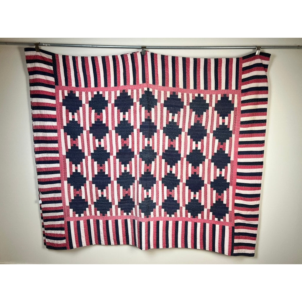 A matching pair of antique American Log Cabin quilts (early 20th century) hangs on the wall, showcasing hand-quilted patriotic red, white, and blue stripes with repeating dark blue squares and pink shapes.