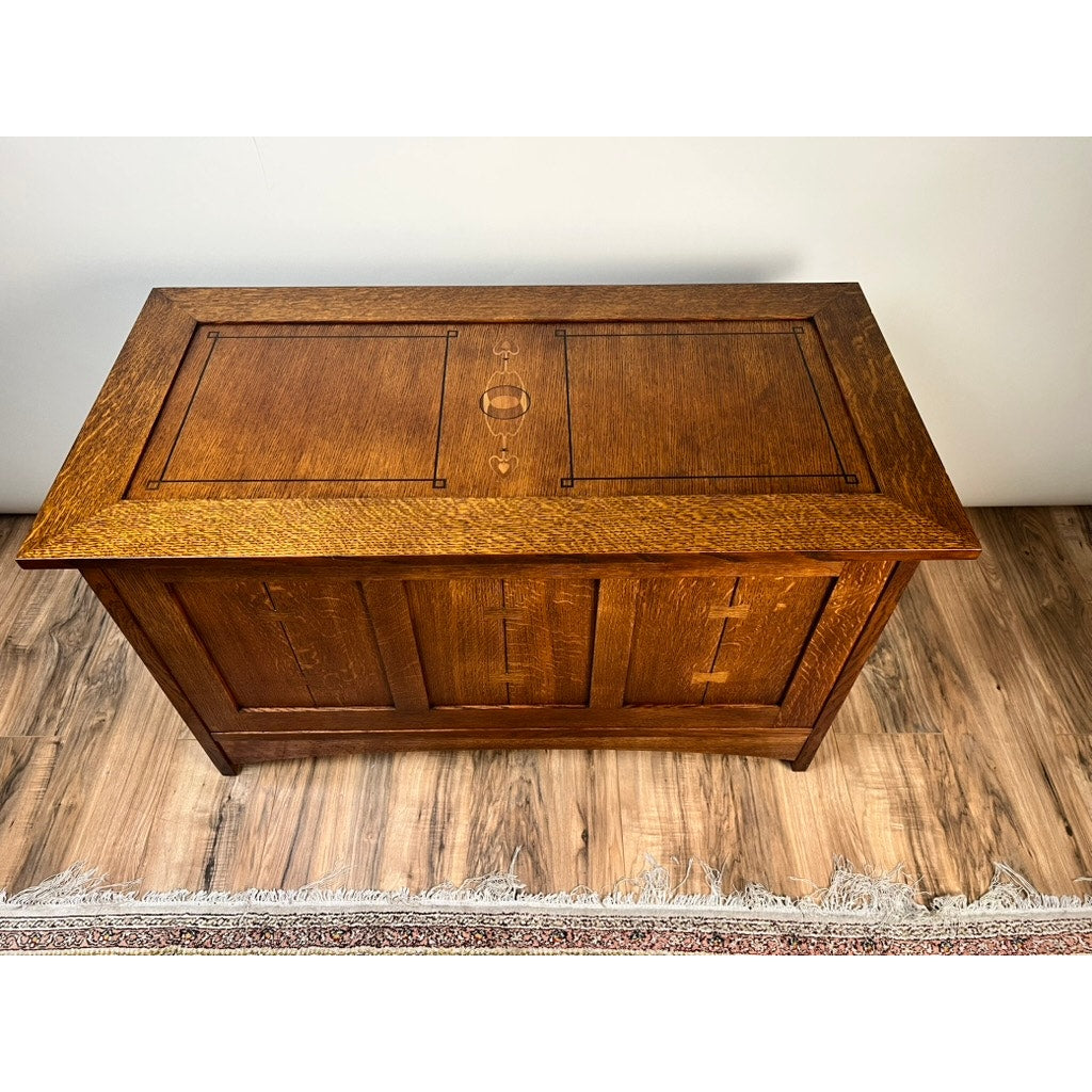 The Vintage Stickley Harvey Ellis Millennium Edition Oak Blanket Chest by Stickley, featuring paneled detailing and decorative inlay, sits on a hardwood floor near a tasseled rug, reflecting classic Arts & Crafts style.
