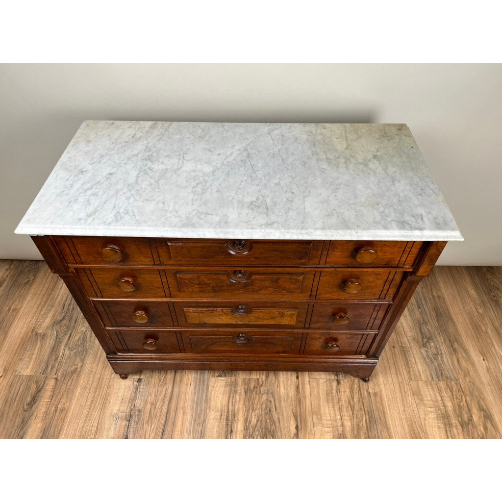 An Antique Victorian Marble Top Four Drawer Chest c. 1910 stands on a wood floor against a plain light-colored wall, featuring a rectangular white marble slab and round wooden knobs.