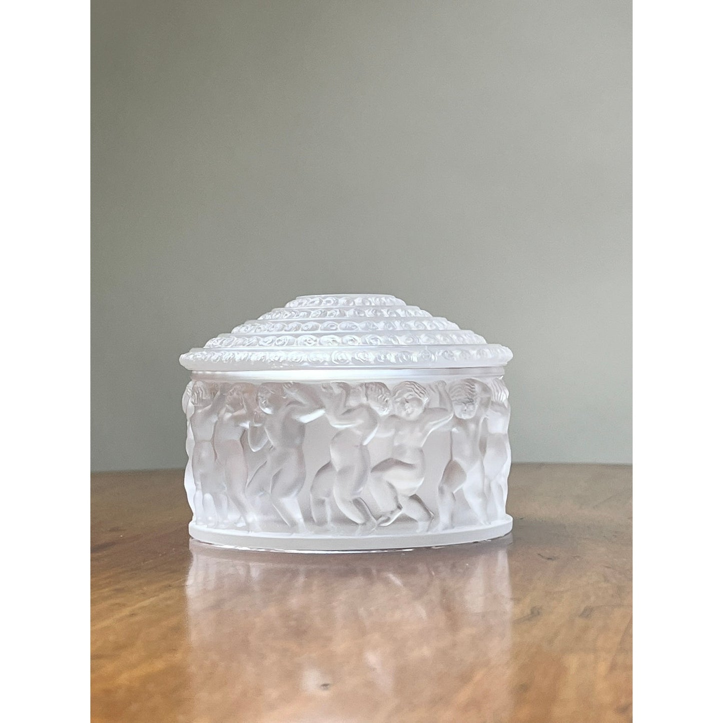 The Vintage Lalique Crystal Enfants Box by Lalique is a white, round decorative container with a dome lid and embossed dancing figures, displayed on a wooden surface against a plain background.