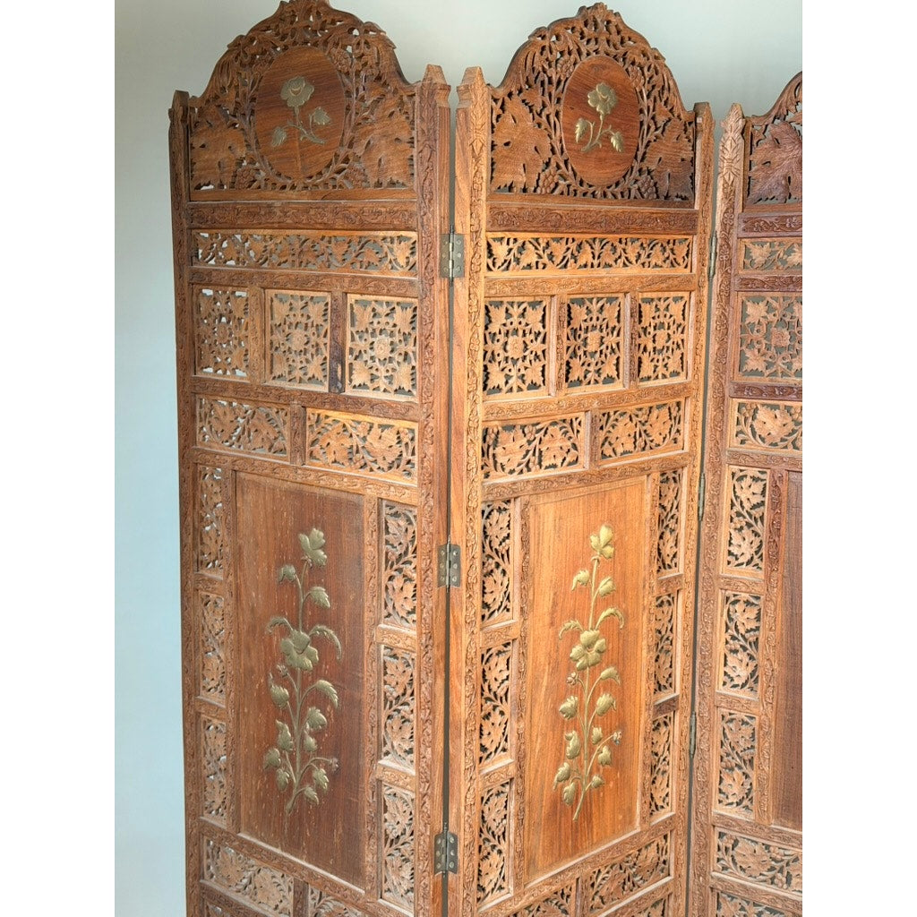 The Early 20th Century Anglo-Indian Carved Teak Four-Panel Screen from Pearl City Antiques features intricately carved wooden panels adorned with applied brass floral decoration and ornate detailing along the top edges.