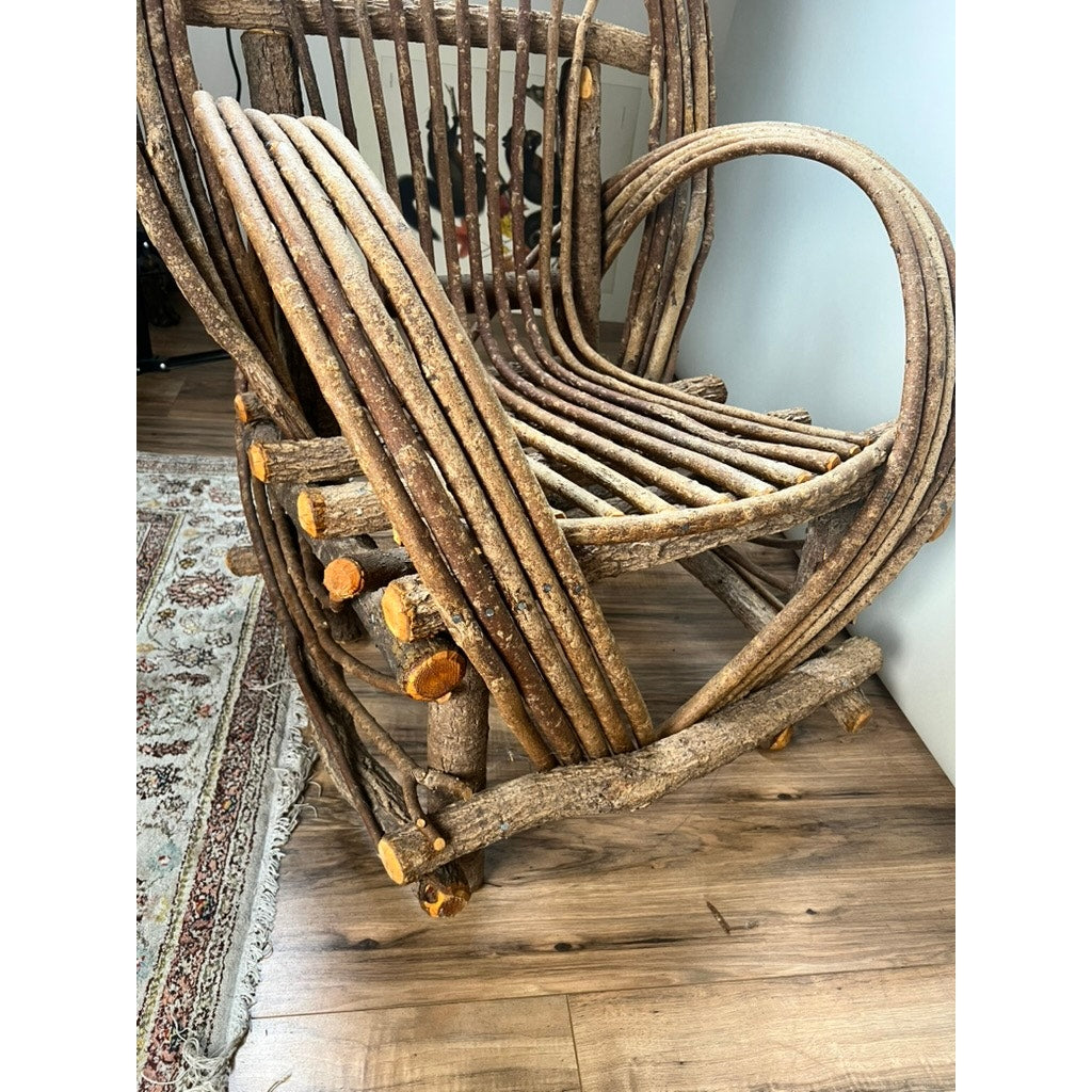 A Vintage Adirondack Bent Willow Arm Chair, crafted from curved, unpolished branches with visible bark and knots, sits on a wooden floor near a patterned rug, showcasing its natural, handcrafted look. Brand: None/Unknown.