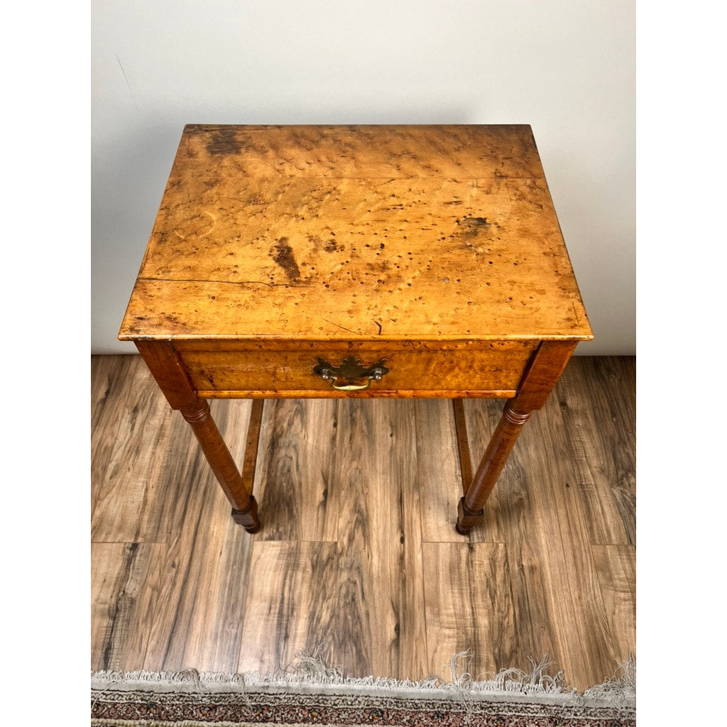 An Antique Sheraton Birdseye Maple One Drawer Stand from the early 19th century with a single drawer, ornate metal handle, turned legs, and a worn honey-colored finish stands on a hardwood floor near a rug. Brand: None/Unknown.