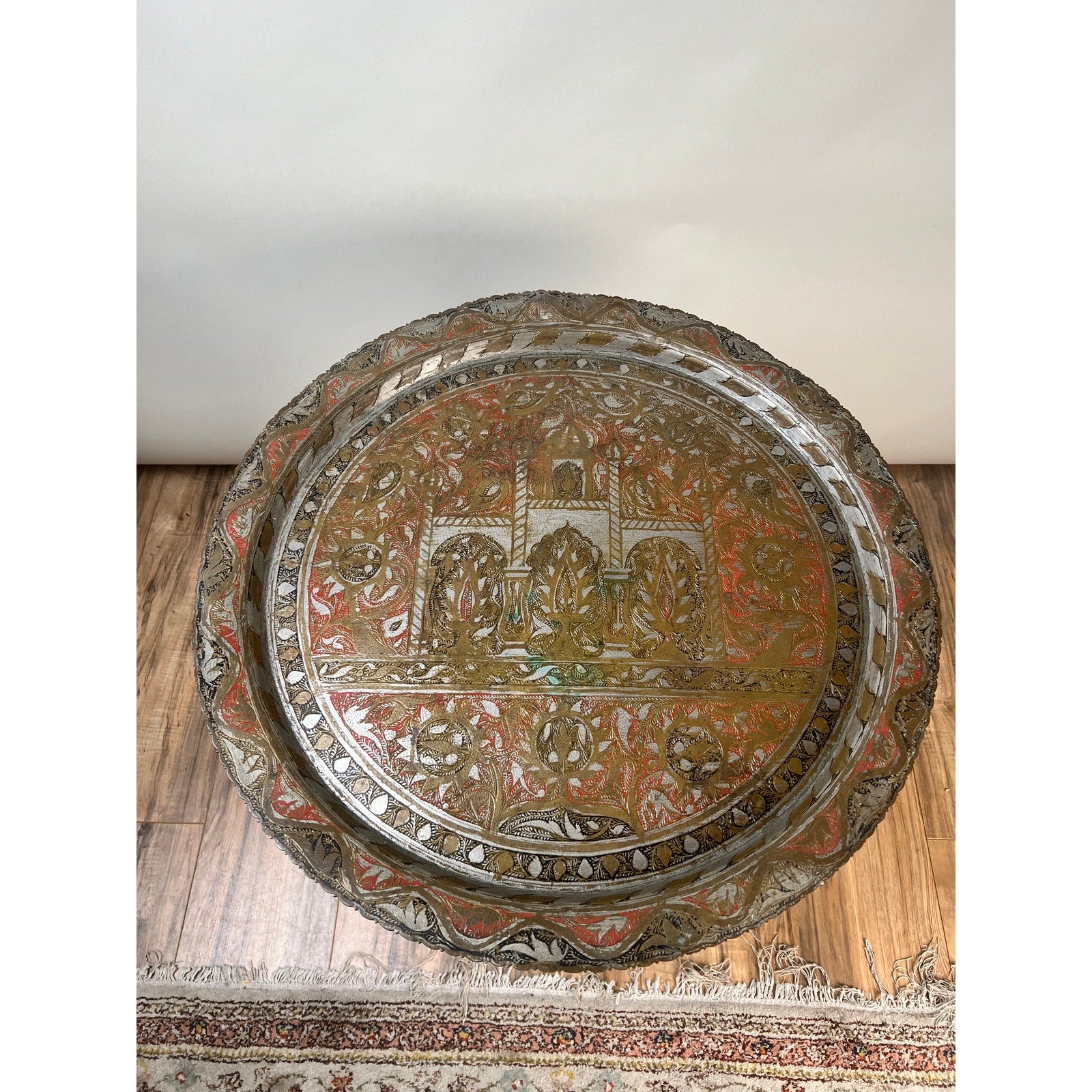 Moroccan or Syrian Moorish folding tray table with a round, hand-chased metal tray showcasing intricate, colorful patterns and engraved architectural designs, displayed on a wooden floor beside a patterned rug.