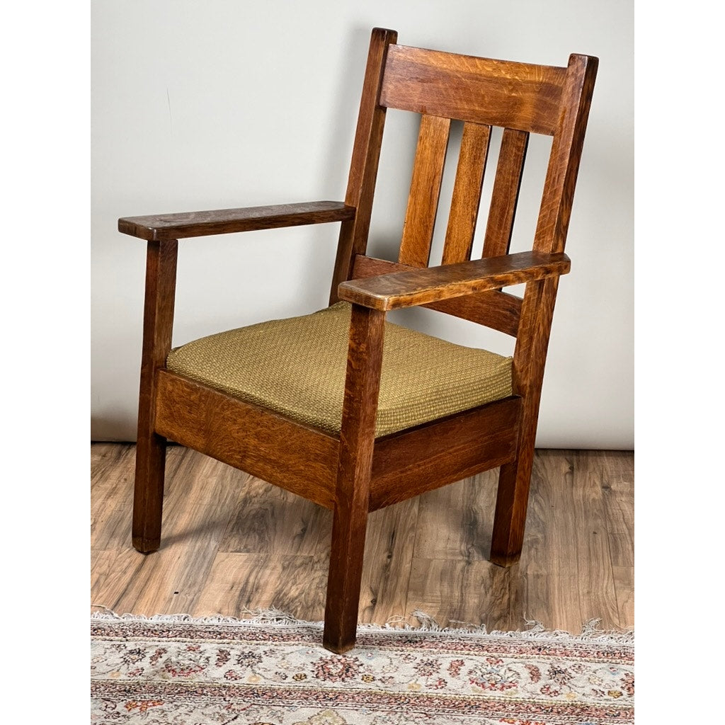 An Antique Quarter Sawn Mission Oak Arm Chair by Quaint Furniture c. 1920 features a slatted backrest and light brown woven seat, set on a wooden floor next to a patterned rug and plain wall, exemplifying Arts & Crafts style.