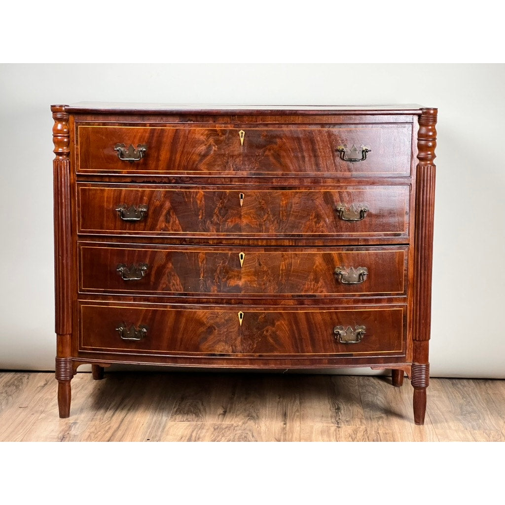 An Antique Sheraton Mahogany Bowfront Chest c. Early 19th Century, with four drawers, brass handles, keyholes, fluted side columns, and tapered legs stands against a light background showcasing its polished wood grain.