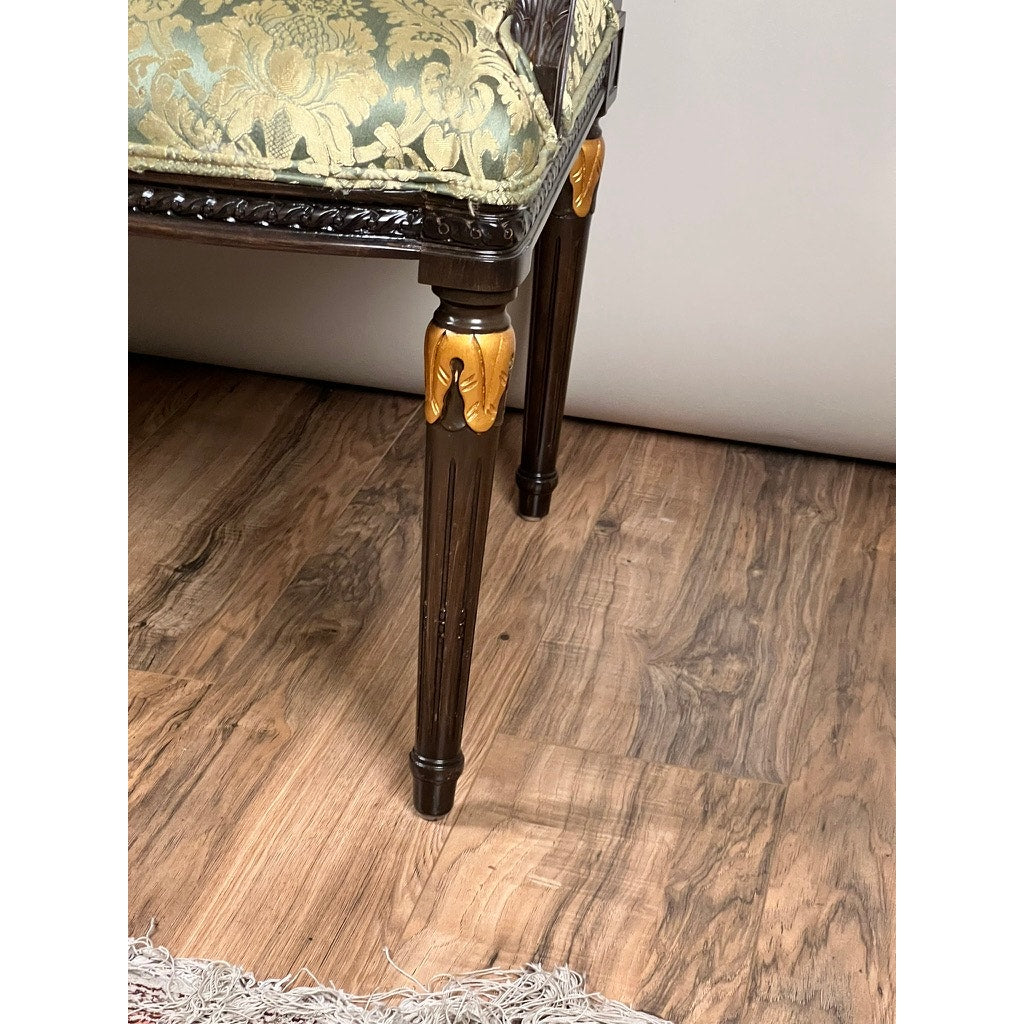A close-up of a vintage Louis XVI style settee in green damask upholstery, featuring an ornate gold-accented wooden leg, shown on a wood floor beside a fringed rug. Brand: None/Unknown.