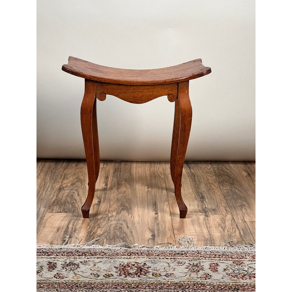 An Antique Arts and Crafts Saddle Shaped Footstool c. 1900 with a curved seat and cabriole legs rests on a wooden floor beside a patterned rug, set against a plain light-colored wall. Brand: None/Unknown.