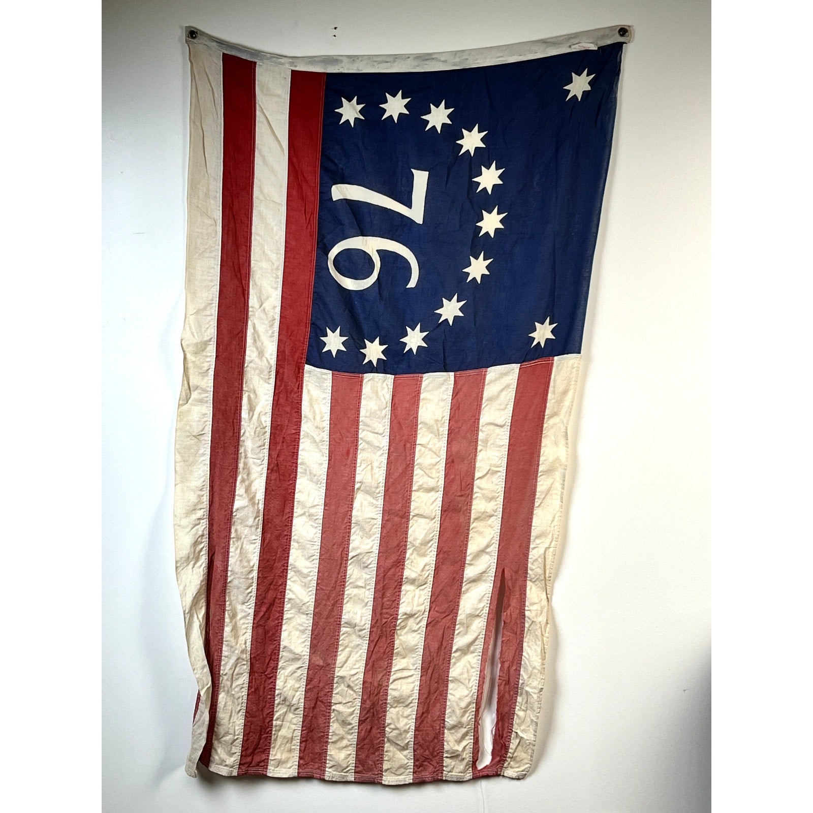 A Vintage Valley Forge “76” Bennington American Flag from the Bicentennial Era (c. 1976), featuring 13 stars in a circle and a blue canton with the number 76 in white at its center, along with classic red and white stripes, is displayed against a plain white wall.