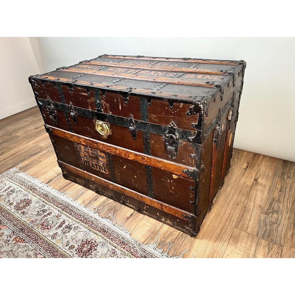 A large CA Taylor Touring Trunk owned by John B. Cordaro, reed player for the Paul Whiteman Orchestra (c. 1930s), sits on a wooden floor near a patterned rug, marked with travel wear and vintage labels.