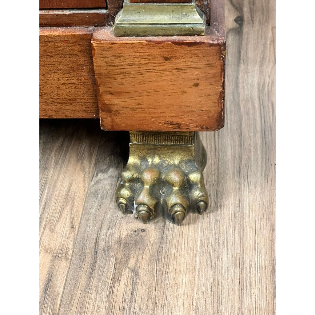Close-up of the Antique French Empire Secrétaire Abattant’s wooden leg, featuring ornate ormolu mounts and a decorative brass claw foot resembling an animal's paw, shown resting on a light brown wood floor.