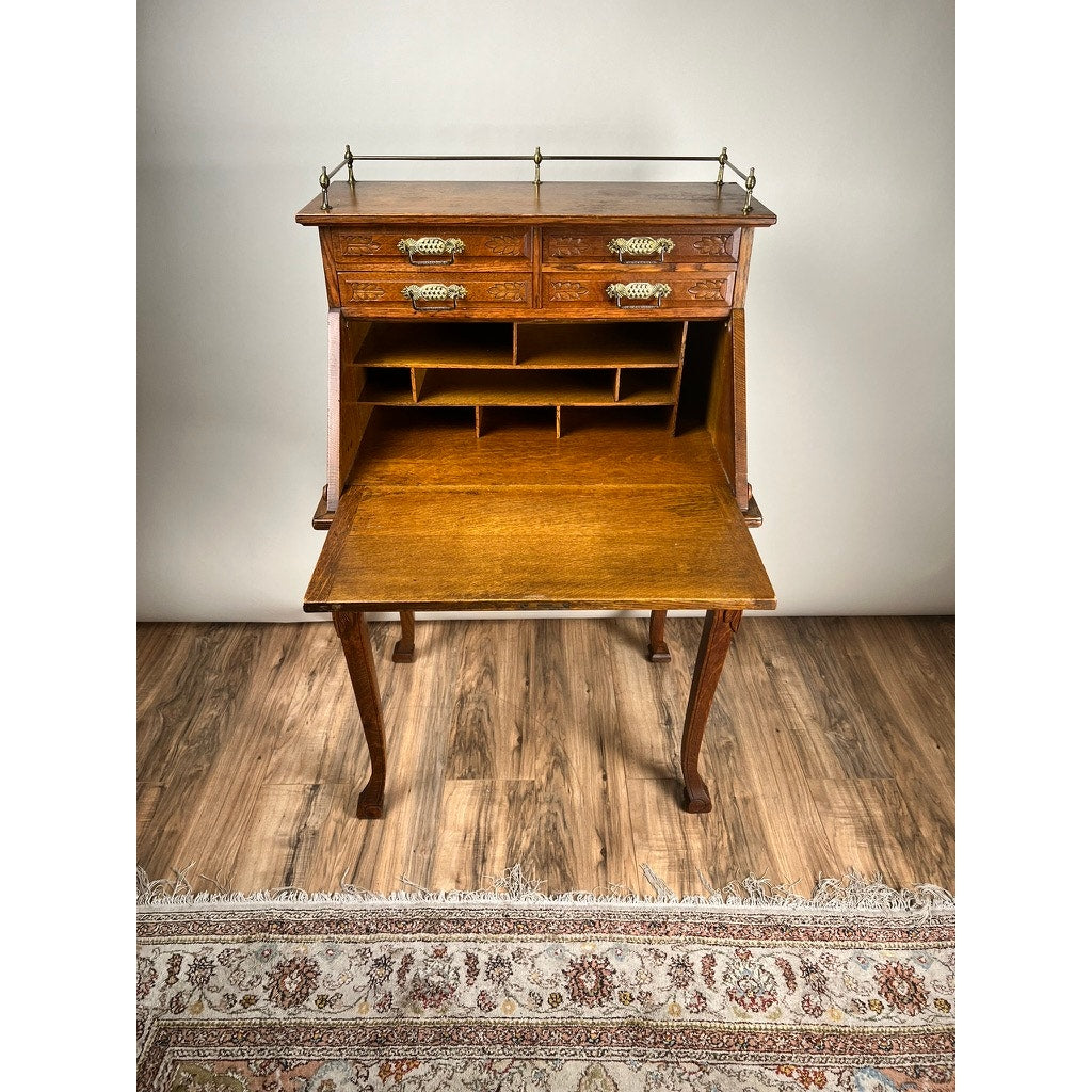 The Antique Victorian Oak Ladies Writing Desk c. 1900 stands on four curved legs with ornate handles and an open fold-down writing surface revealing compartments, set on a wooden floor with a patterned rug in front.