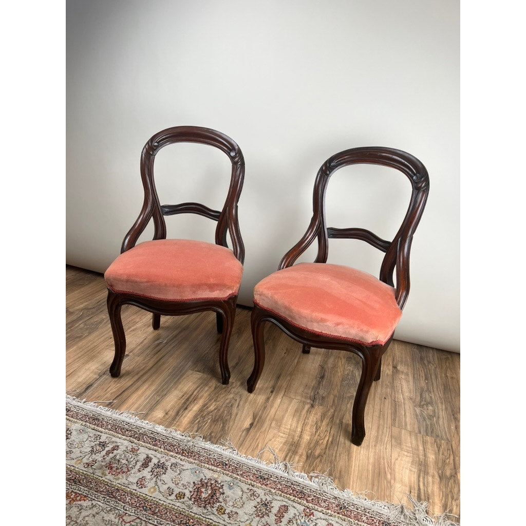 An antique pair of Victorian walnut balloon back side chairs with reddish-orange velvet seats are set side by side on a wooden floor next to a patterned rug, against a plain light-colored wall.