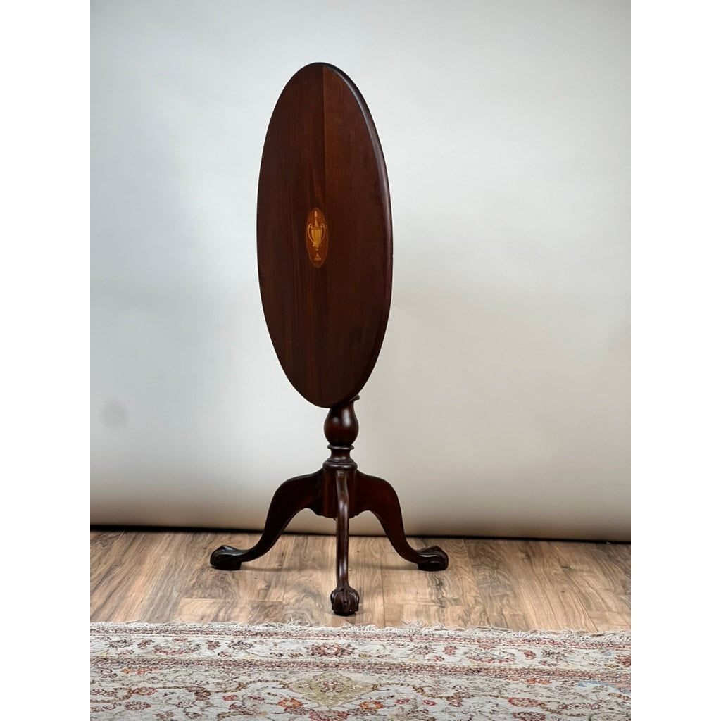 The Antique George III Tilt-top Tea Table by Pearl City Antiques, dating to the early 19th century, features a dark wood oval top, pedestal base, and three elegantly curved legs. It is displayed on a wooden floor beside a patterned rug, set against a plain light background.