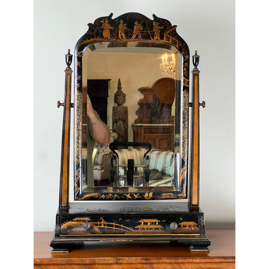A Fine Antique Chinoiserie Black Lacquer Dressing Mirror c. Late 19th Century from Pearl City Antiques, featuring Asian-inspired gold detailing, stands on a wooden table. The mirror reflects part of a room with furniture, a statue, and a chandelier, along with a person’s arm holding a phone.